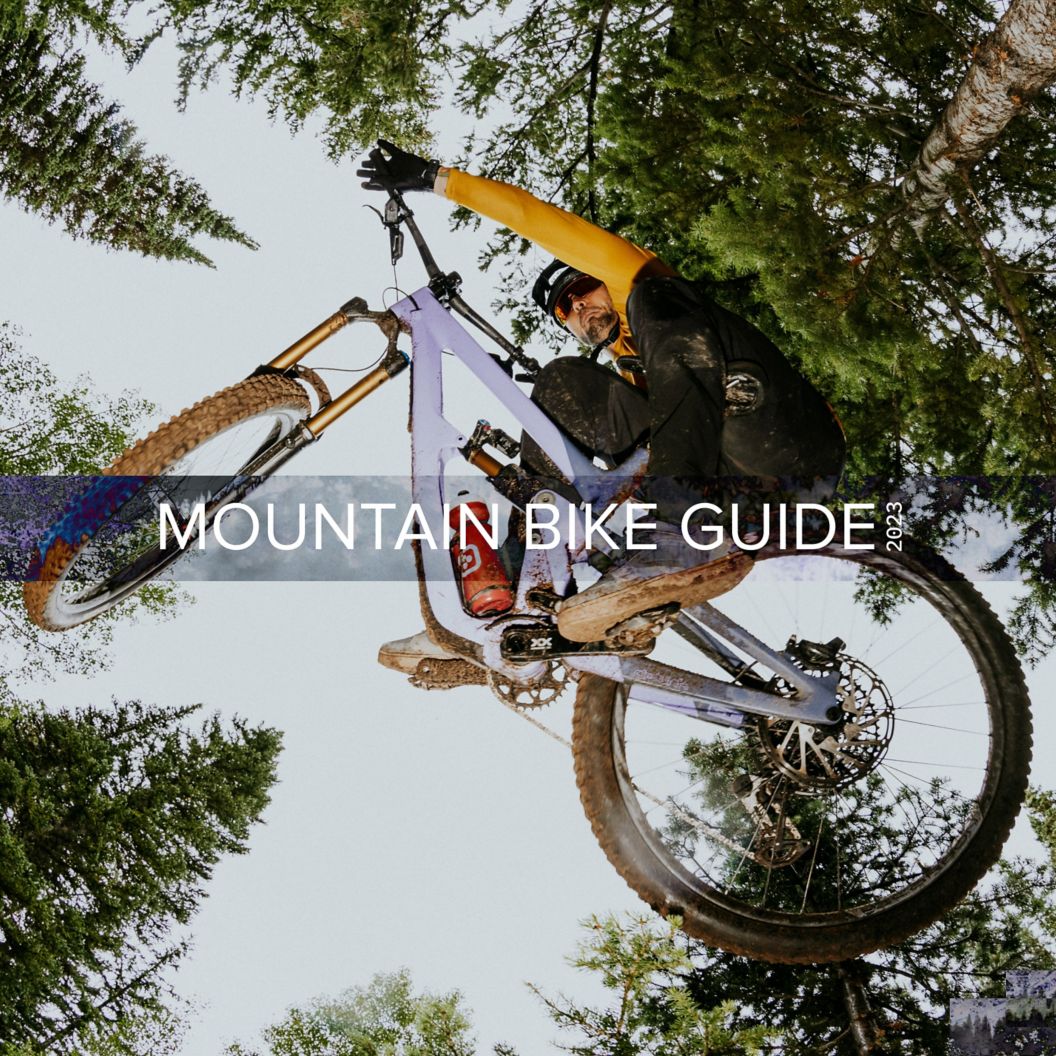     2023 Mountain Bike Guide text on an image looking up at an MTB rider in fall apparel table-topping in an alpine tree forest. 